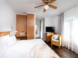 Una habitación de hotel con cama, televisor sobre un mueble de madera, una silla de mimbre, ventilador de techo y armario.