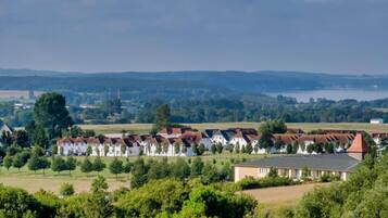 Une rangée de maisons blanches aux toits rouges, entourées d’arbres et de champs verdoyants, avec une étendue d’eau et des collines en arrière-plan.