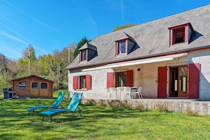 Ein zweistöckiges Haus mit Terrasse, eine kleine Holzhütte und zwei blaue Lounge-Sessel.