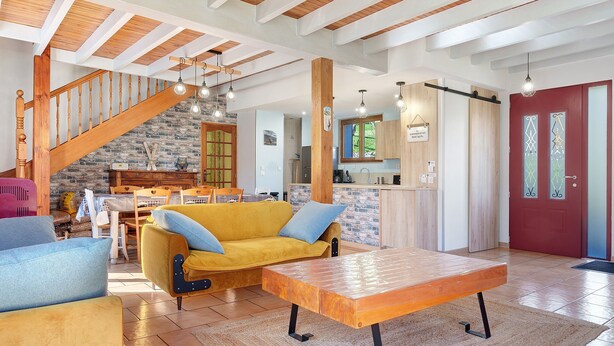 A spacious living area with a wooden staircase, a yellow sofa, a wooden coffee table, and a kitchen area with wooden cabinets and a red door. - Holiday Home 'Gite 8 Personnes, Val D'azun' with Mountain View, Private Terrace and Wi-Fi (Bun)