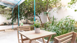 Una mesa y sillas de madera bajo un toldo azul, rodeadas de macetas con plantas y un árbol.