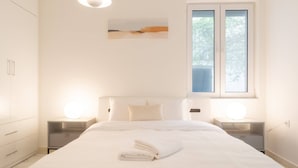 Una cama bien hecha con una toalla doblada, una lámpara de noche y una ventana con persianas.