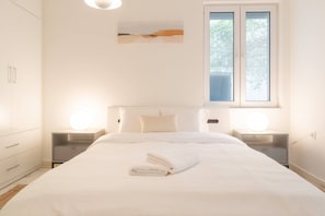 A neatly made bed with a folded towel, a bedside lamp, and a window with blinds. - A Haven in Kypseli A Basement Apartment (Athens)