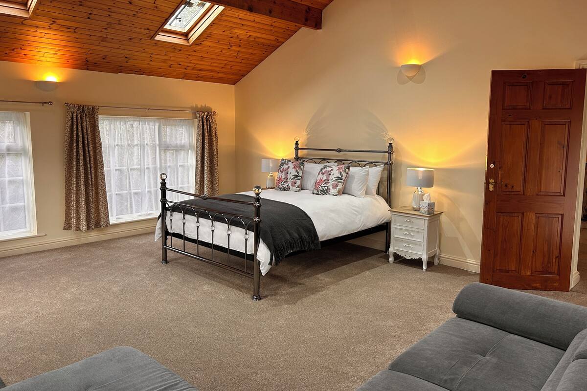 A bedroom with a four-poster bed, a nightstand with a lamp, a wooden door, and a window with curtains.