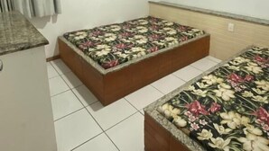 Una habitación con dos camas, ropa de cama estampada con motivos florales, ventilador de techo, ventana con cortinas y suelo de baldosas.