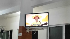Un área de comedor con un televisor en la pared mostrando un personaje de caricatura. Hay una mesa con sillas, un pilar de ladrillo y una ventana con persianas.