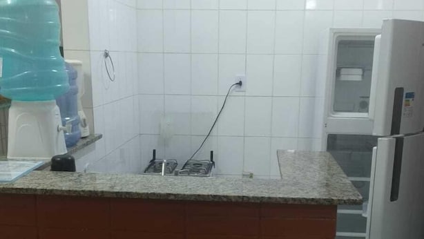 Una cocina con paredes de azulejos blancos, encimera de granito, dos taburetes de madera, un refrigerador de acero inoxidable y un dispensador de agua.