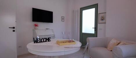 A modern living room with a white sofa, a round table with a 'WELCOME' sign, a flat-screen TV, and a green-framed door.
