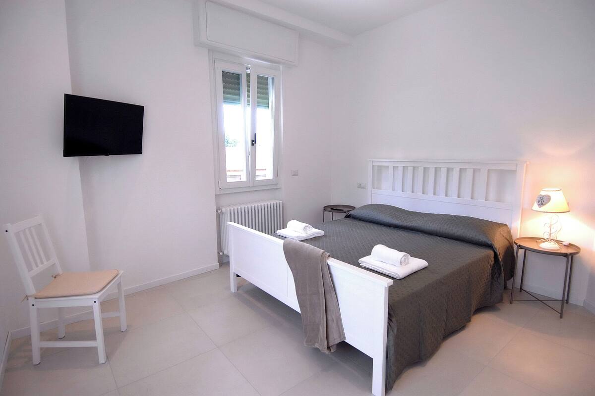 A bedroom with a bed, a chair, a bedside table, a lamp, and a TV mounted on the wall.