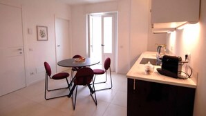 A compact kitchen with a dining table and chairs, a countertop with a coffee machine, and a door leading to a bright room. - Le Case di Elena (Gignese)