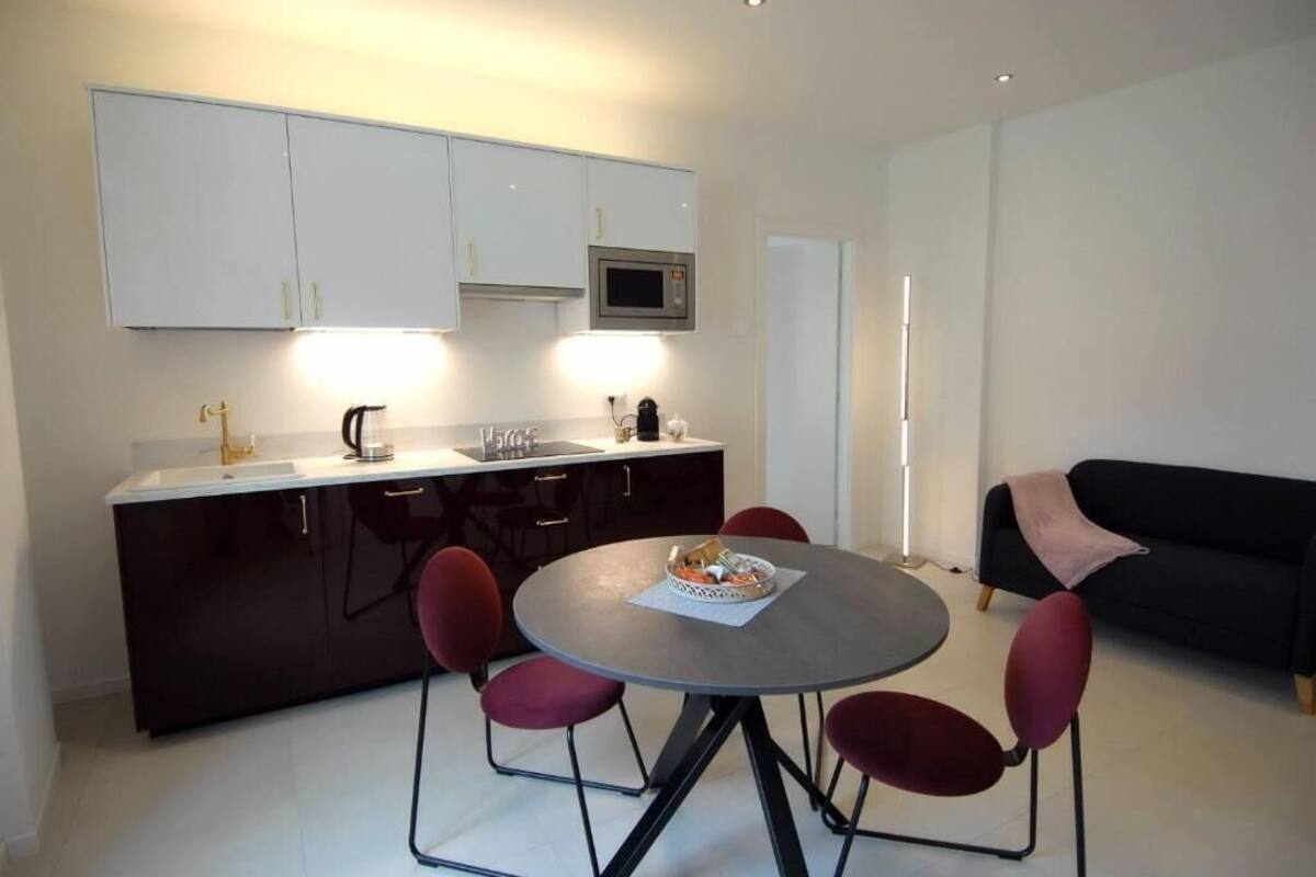 A compact kitchen with white cabinets, a microwave, and a sink. There is a dining table with four chairs and a small round table with a basket. A black sofa is visible in the background.