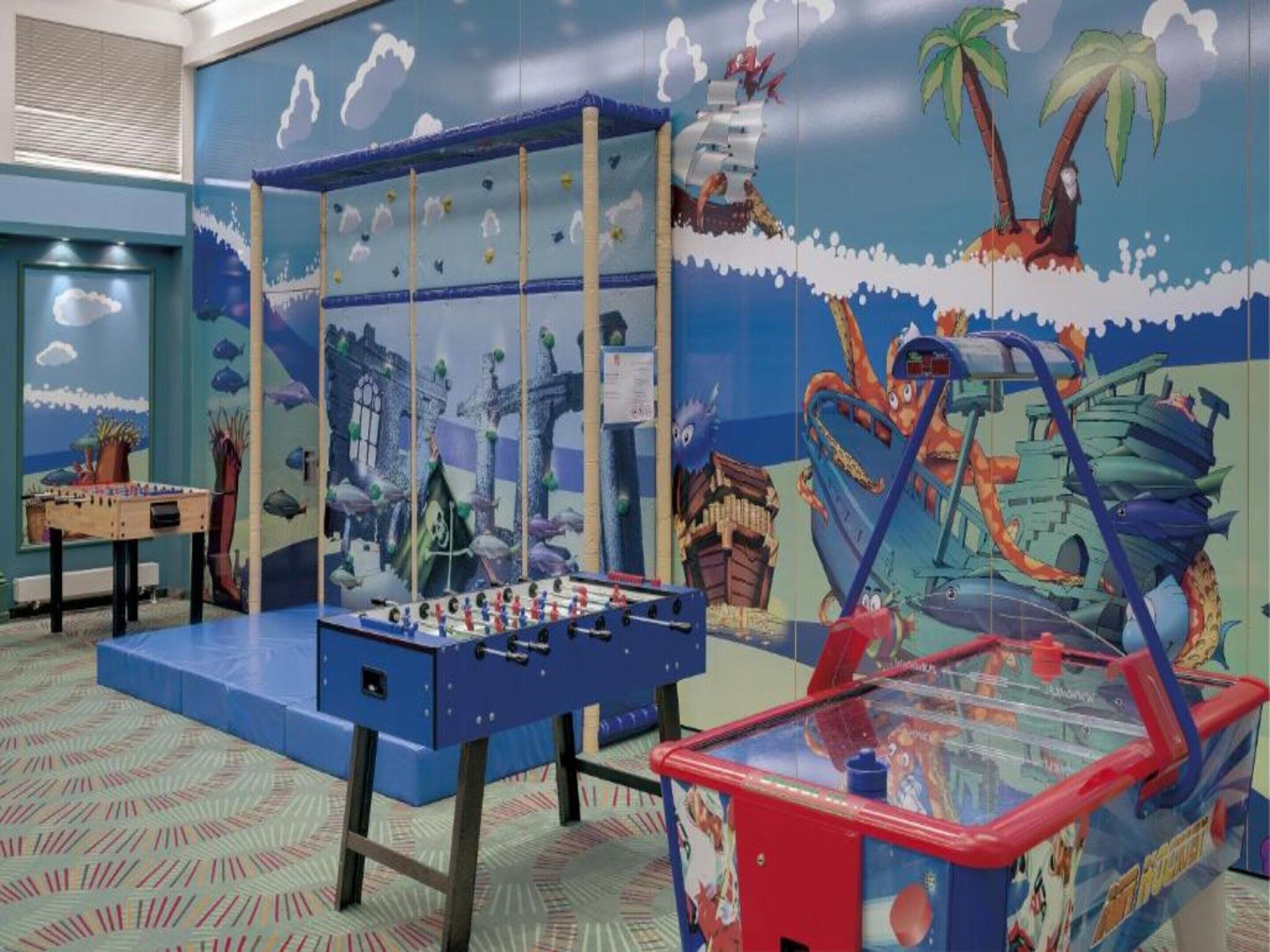 Una habitación con mesa de futbolín, mesa de tenis de mesa y un mural que muestra una escena tropical con palmeras y un barco.