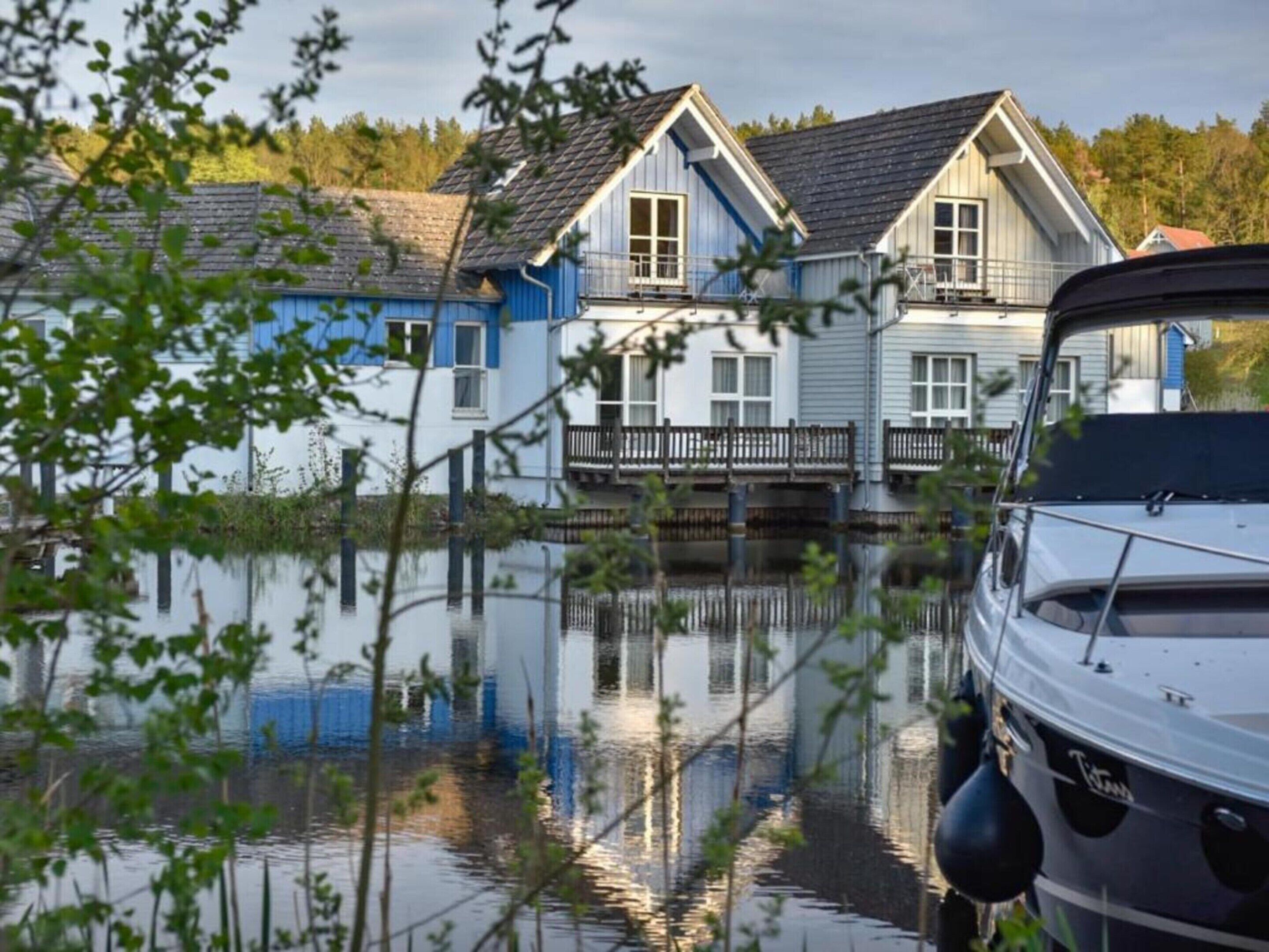 A houseboat docked at a calm body of water with a clear reflection, surrounded by greenery.