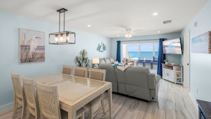 A modern living room with a dining area, a ceiling fan, a large window with a view, and a flat-screen TV. - Coastal Comfort with Gulf-Front Views (Sarasota)