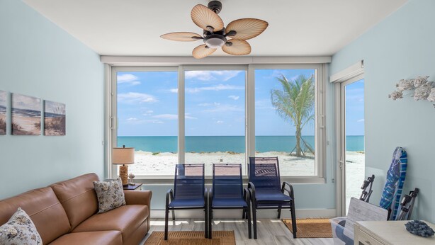 A room with a large window, a ceiling fan, a brown leather sofa, and a table with chairs. - Coastal Comfort with Gulf-Front Views (Sarasota)
