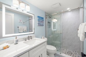 A bathroom with a large mirror, two sinks, a toilet, and a glass-enclosed shower. - Coastal Comfort with Gulf-Front Views (Sarasota)