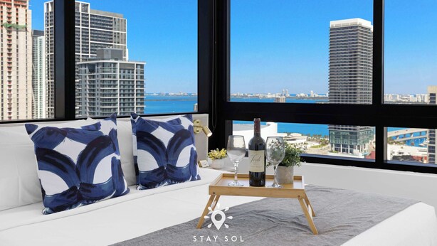 A modern hotel room with a large window offering a city view, a sofa with decorative pillows, a tray with a wine bottle and glasses, and a small plant. - Free Parking | Miami Gem | Smart 2BR Corner View (Miami)