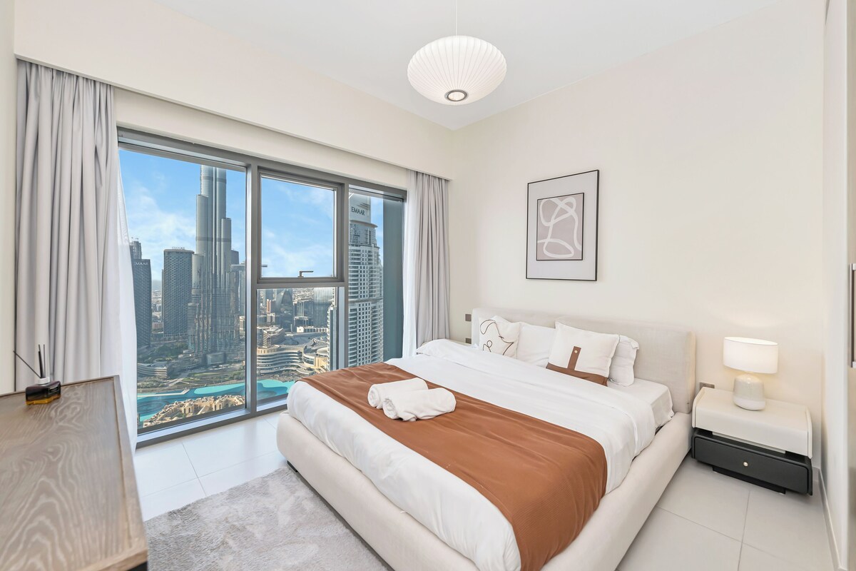 Ein modernes Hotelzimmer mit einem großen Bett, einem Nachttisch mit Lampe, einem gerahmten abstrakten Kunstwerk und Blick auf die Stadt durch raumhohe Fenster.