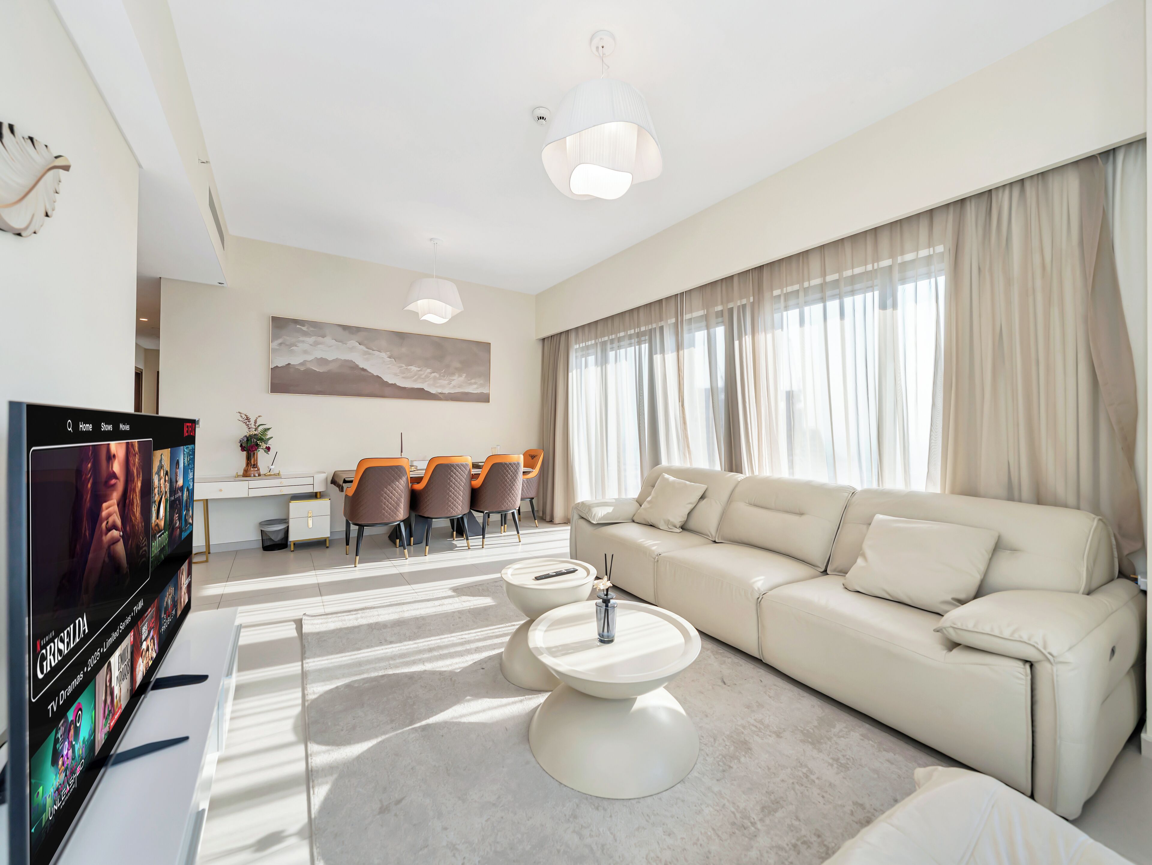 A modern living room with a sofa, coffee table, and TV. There is a dining area with chairs and a table, and a large window with curtains.