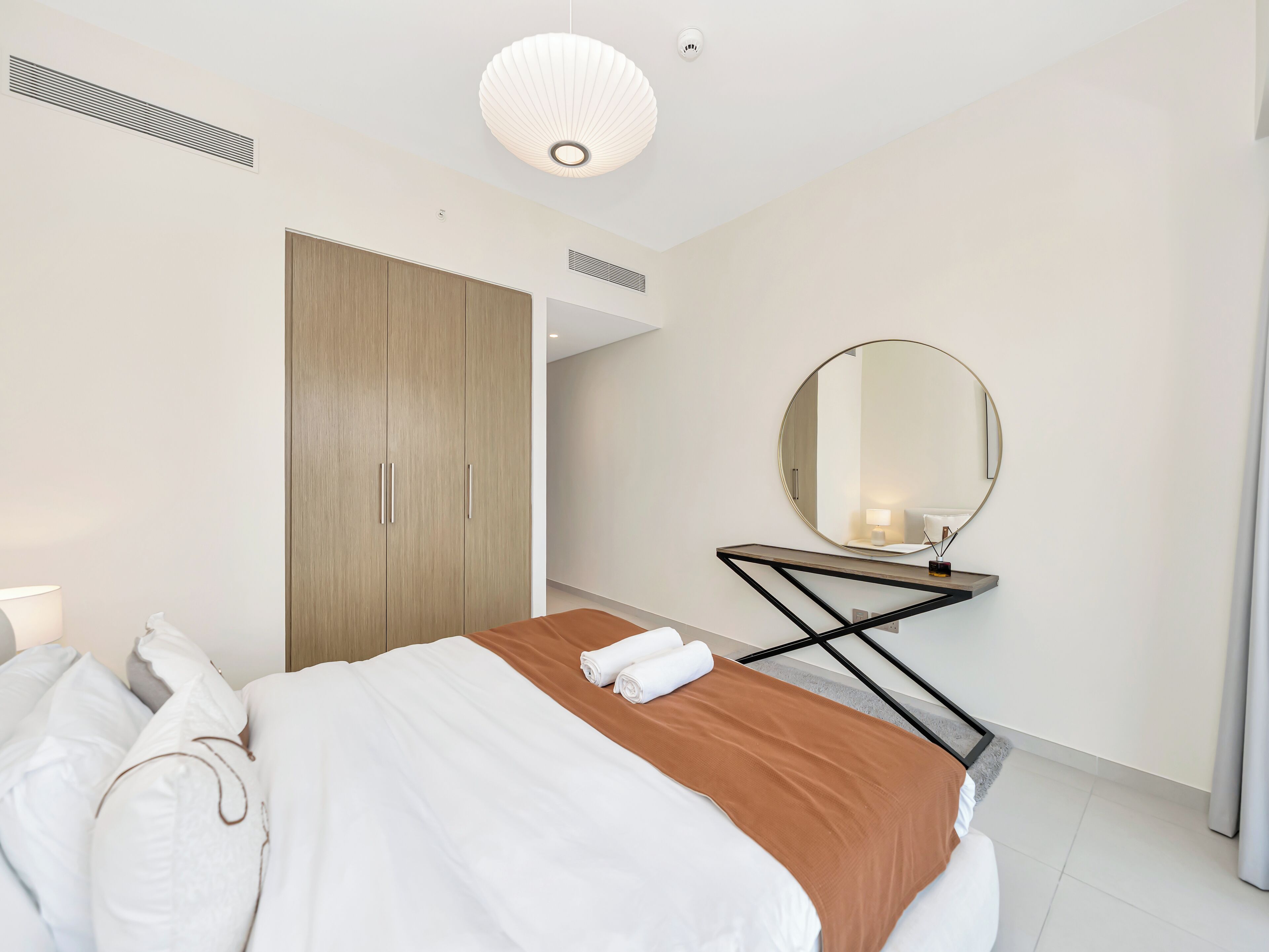 A modern bedroom with a bed, a wooden wardrobe, a round mirror, and a bedside table.