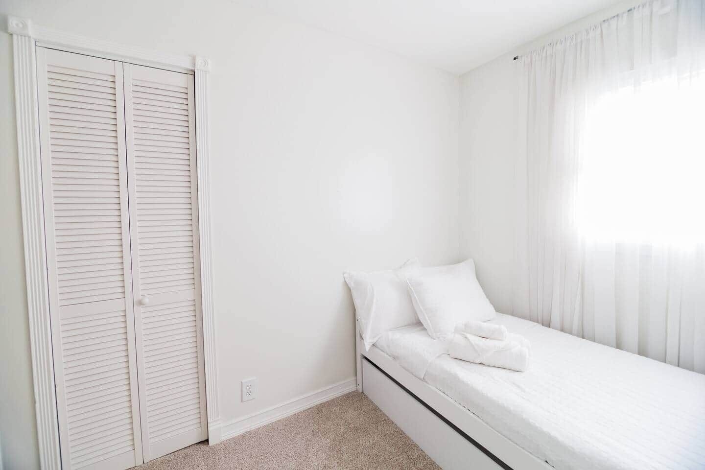 Ein kleines, weißes Schlafzimmer mit einem Einzelbett, einem Kleiderschrank und einem Fenster mit durchsichtigen Vorhängen.