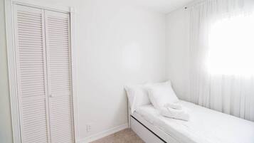 Ein kleines, weißes Schlafzimmer mit einem Einzelbett, einem Kleiderschrank und einem Fenster mit durchsichtigen Vorhängen.