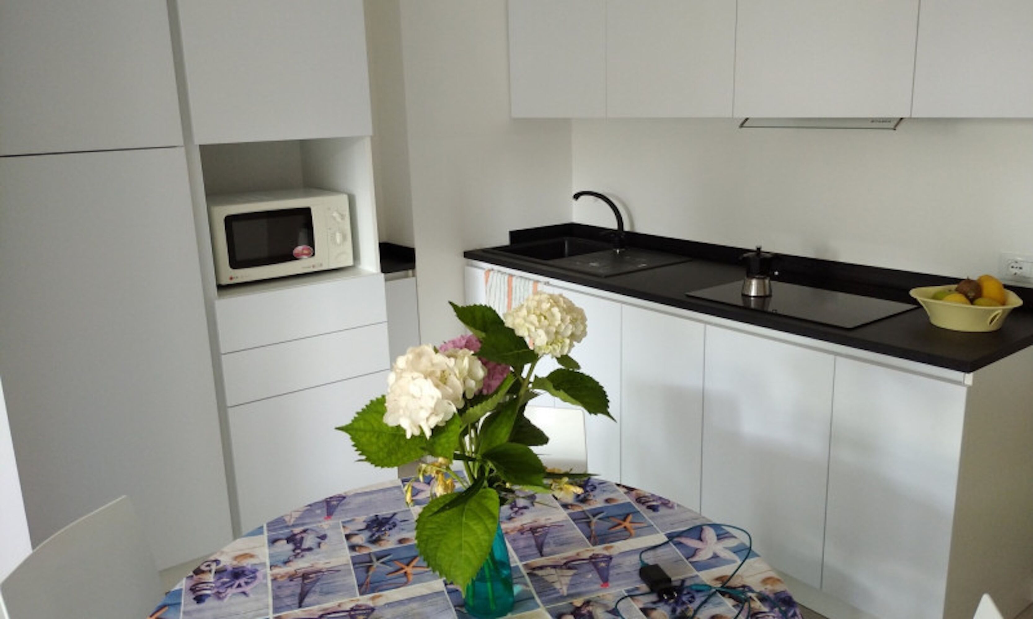 A compact kitchen with a microwave, sink, and a small fruit bowl. A vase with flowers is on a table with a nautical-themed tablecloth.