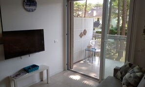A living room with a flat-screen TV, a small table with a blue basket, a sofa with patterned cushions, and a sliding glass door leading to an outdoor area with a white fence and a chair. - Flat on the Shores of Lignano (Lignano Sabbiadoro)
