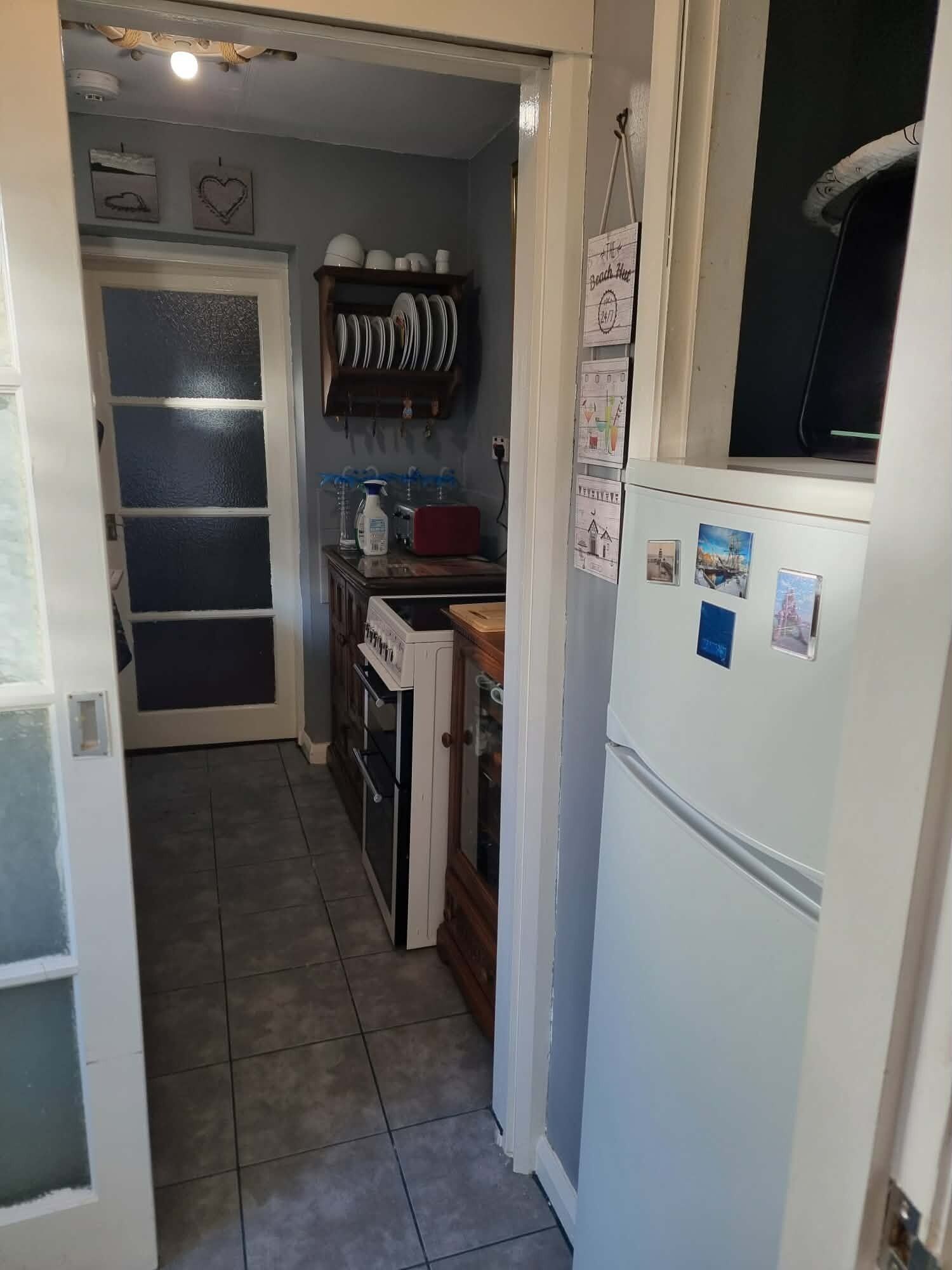 A narrow kitchen with a refrigerator, oven, and a shelf with dishes.