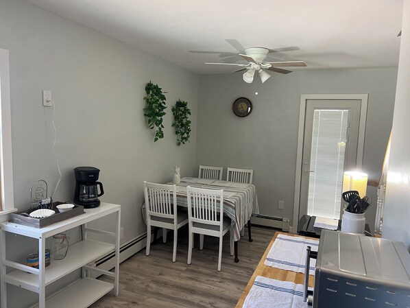 A compact kitchen with white cabinetry, a coffee maker, a dining table with chairs, a ceiling fan, and a clock on the wall.