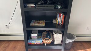 A bookshelf with video games, books, and a Scrabble box. - Cozy 3BR Apartment in Peaceful Neighborhood With Kitchenette (Commack)