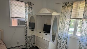 A bedroom with a bed, a vanity with a mirror, a television, and a window with blinds. - Cozy 3BR Apartment in Peaceful Neighborhood With Kitchenette (Commack)
