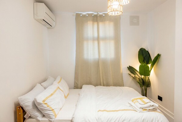Una cama bien tendida con sábanas blancas y detalles dorados, un aire acondicionado de pared, una ventana con cortinas color beige y una planta en maceta a la derecha.