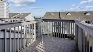 Een houten terras met een metalen reling, dat leidt naar een gebouw met meerdere daken en een heldere lucht.