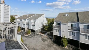 Een rij moderne woongebouwen met twee verdiepingen, balkons, een parkeerplaats en een helderblauwe lucht.