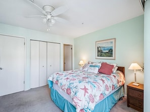 Une chambre à coucher avec un lit, un ventilateur de plafond, une table de chevet, une lampe et un tableau encadré au mur.
