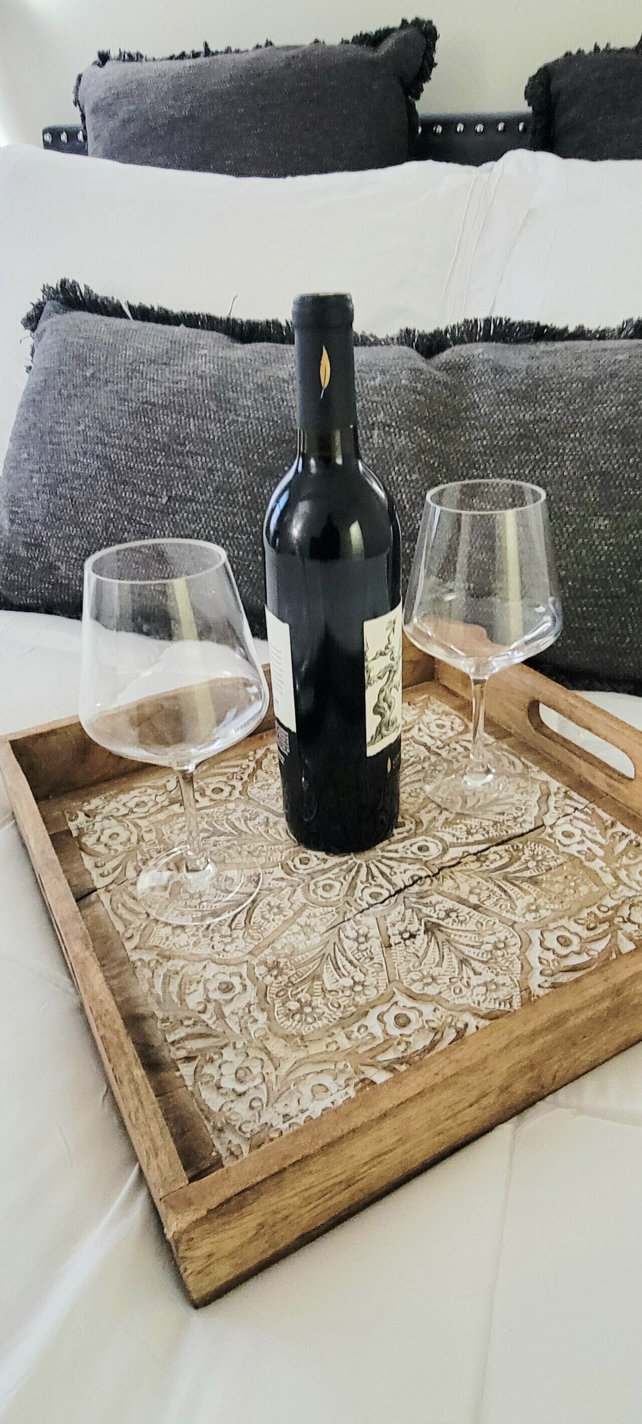 A bottle of wine on a tray with two glasses on a bed with patterned pillows.