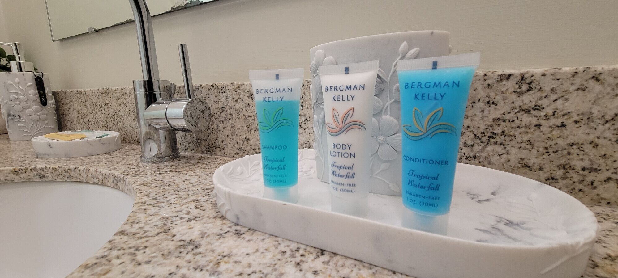 Et badeværelsesbord med en marmorvask, et spejl og tre Bergman Kelly produkter: shampoo, body lotion og balsam.