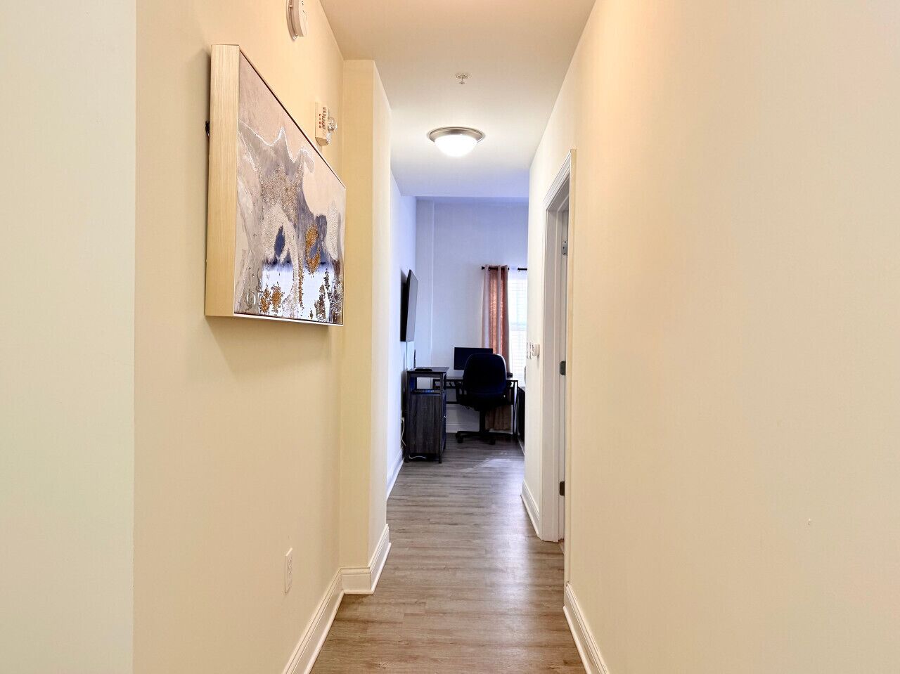 A hallway with a wooden floor, a painting on the left wall, and a desk with a chair in the background.