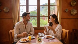 Un couple prend son petit-déjeuner à une table en bois, avec vue sur le paysage par la fenêtre et des assiettes décoratives accrochées au mur.