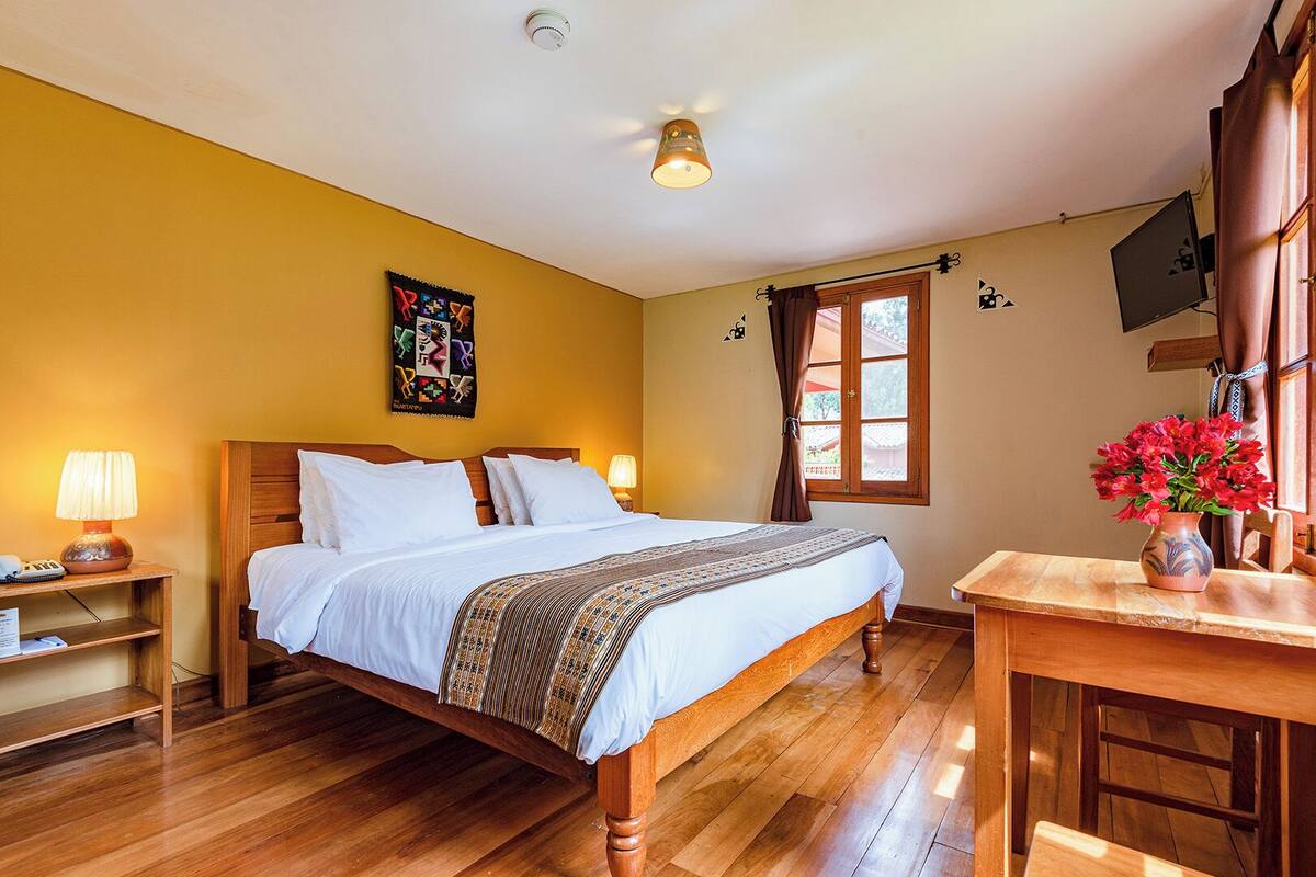 Un dormitorio con una cama de madera, una mesita de noche con una lámpara, un escritorio con un florero y una ventana con cortinas.