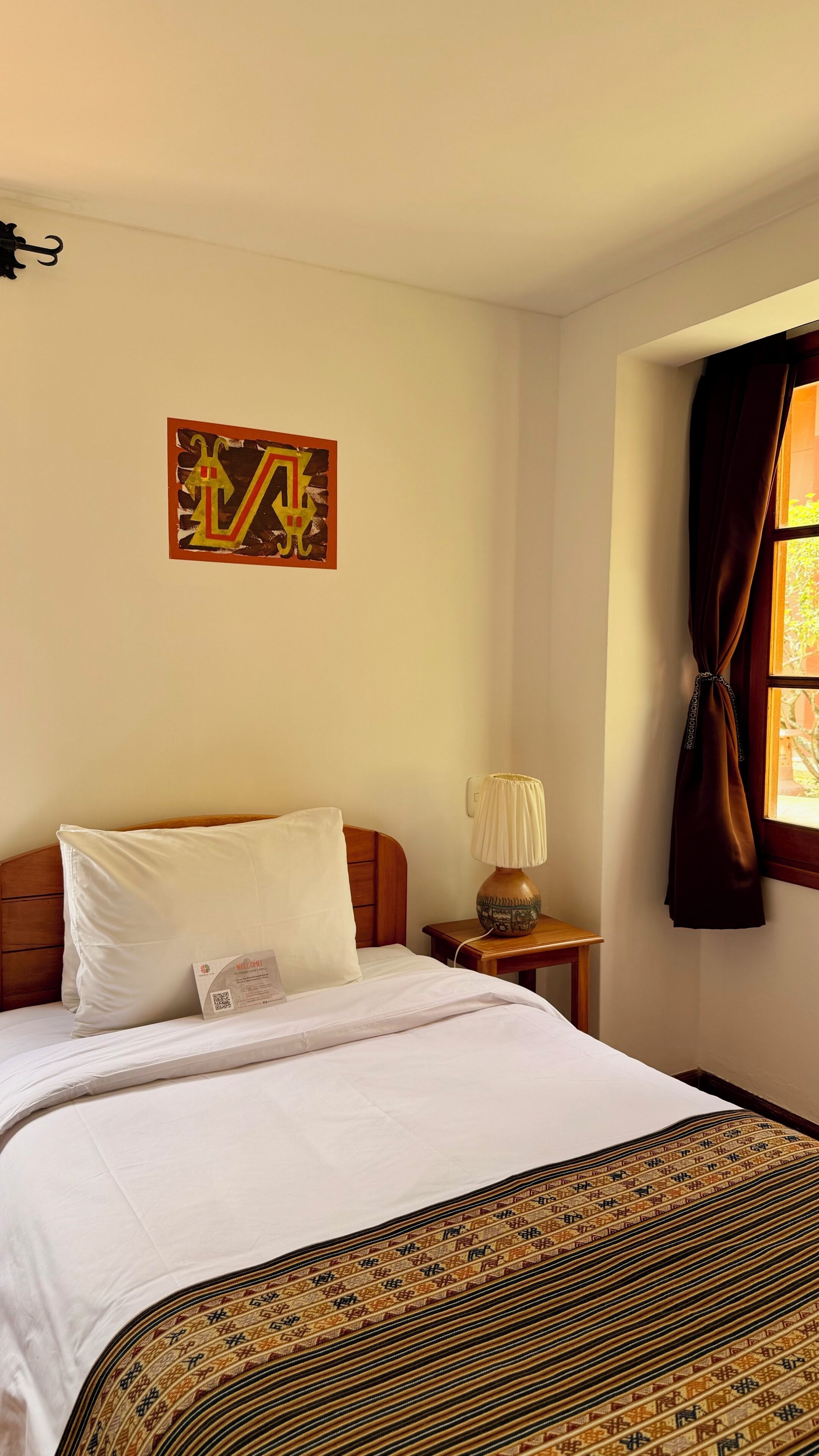 A hotel room with a bed, a bedside table with a lamp, a window with curtains, and a wall decoration.