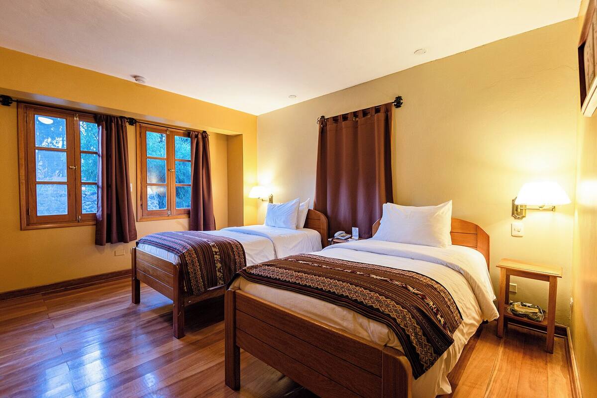 Una habitación de hotel con dos camas de madera, piso de madera y un ventanal con cortinas.