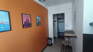 Een hal met oranje muren, een klein zithoekje met krukken, en een deur naar een kamer met een bed en een kledingkast.