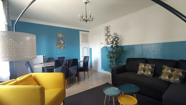 Een moderne woonkamer met een zwarte bank, een geel fauteuil en bijzettafeltjes in blauw en geel. Er is een eethoek met een witte tafel en blauwe stoelen.