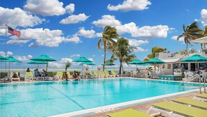Une piscine entourée de palmiers, avec des chaises longues et des parasols, sous un ciel dégagé.