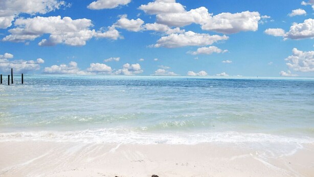 Une plage à l’eau claire, avec un rivage sablonneux et un ciel bleu parsemé de nuages.