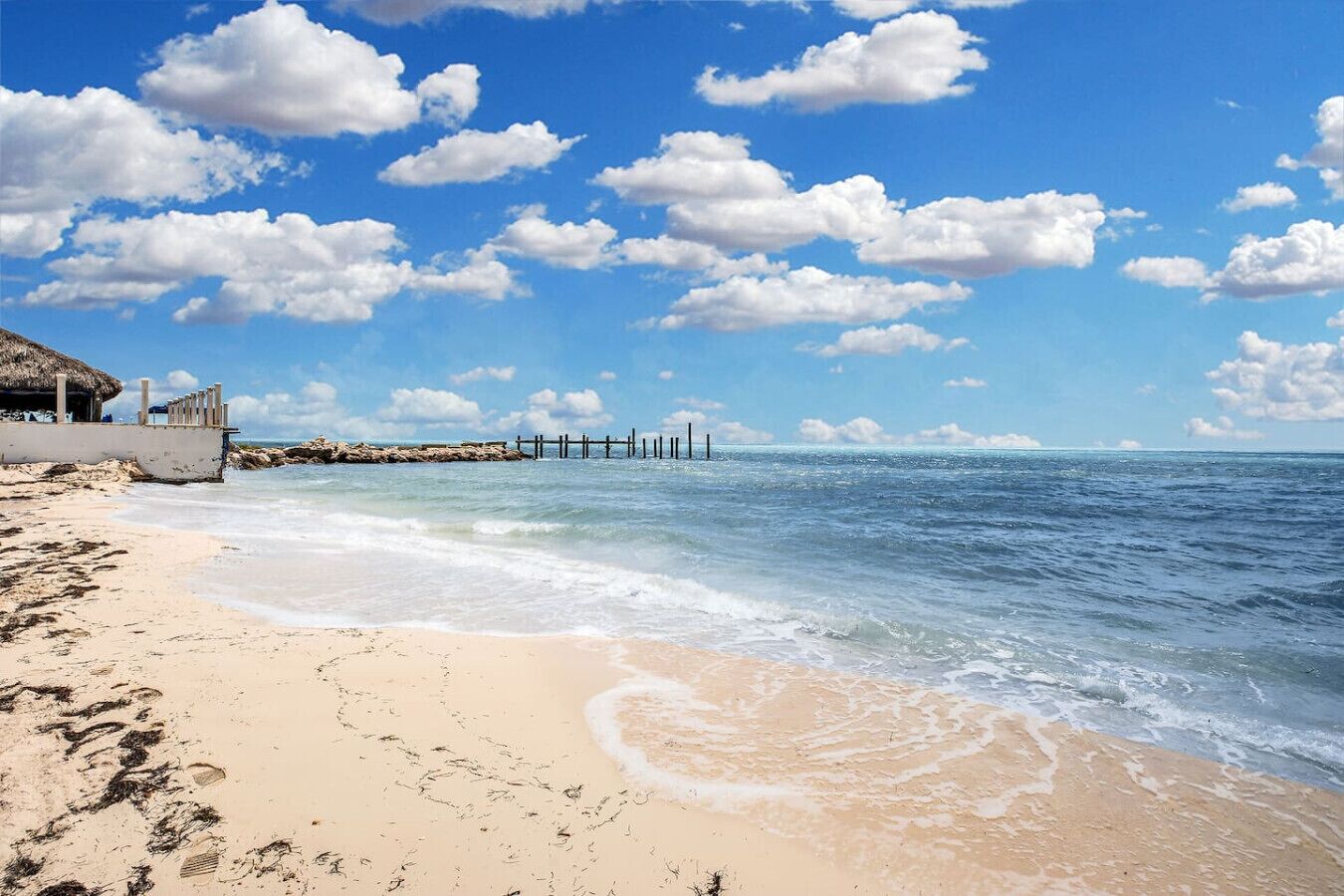 A beach with a thatched-roof structure, a wooden pier, and clear blue skies.