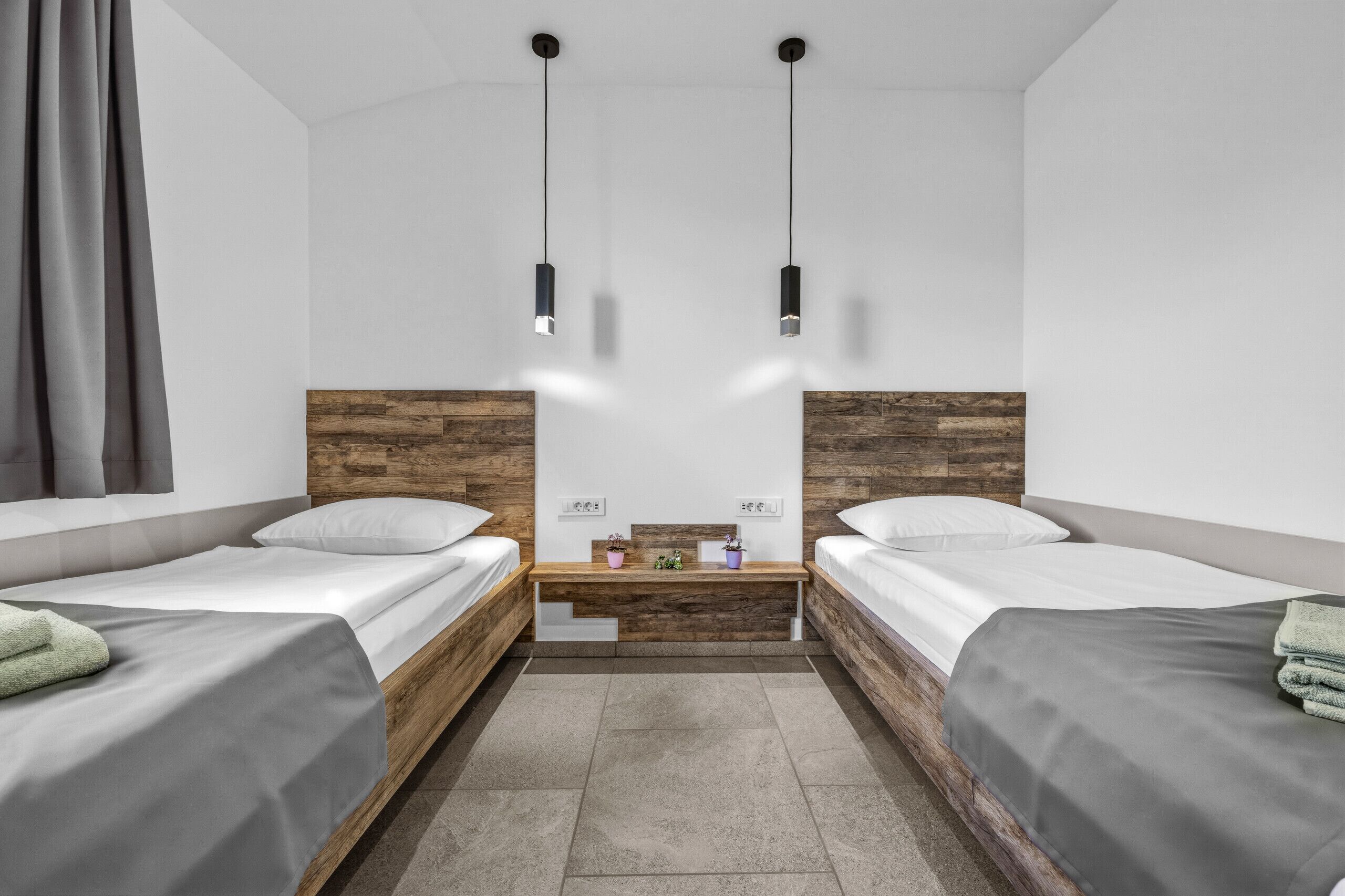 A modern hotel room with two single beds, a wooden headboard, a small bench, and pendant lights.