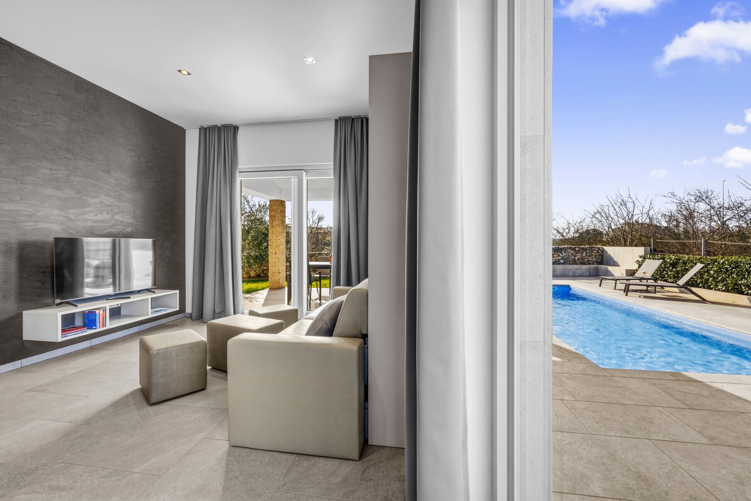 A modern living room with a flat-screen TV, a sofa, and a poolside area with lounge chairs.